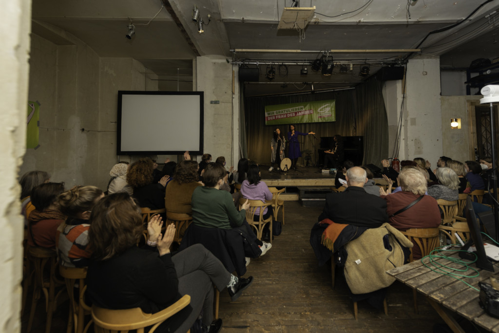 Blick über den Veranstaltungsraum von hinten: Besucher:innen auf Stühlen und Bühne vorne, auf der gerade die Musiker:innen Sakîna Têyna, Basma Jabr und Mahan Mirarab sind