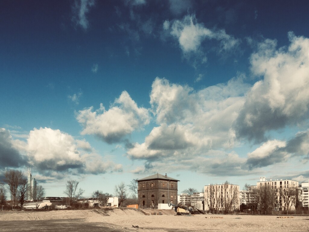 Der Wasserturm in der Freien Mitte, 2019: Rund um den braunen Quader ist noch freie Fläche, und zwar Sand, ein paar Bäume im weitern Umfeld; im Hintergrund sind Häuser zu sehen, darüber ein Himmel mit malerischen Wolken.