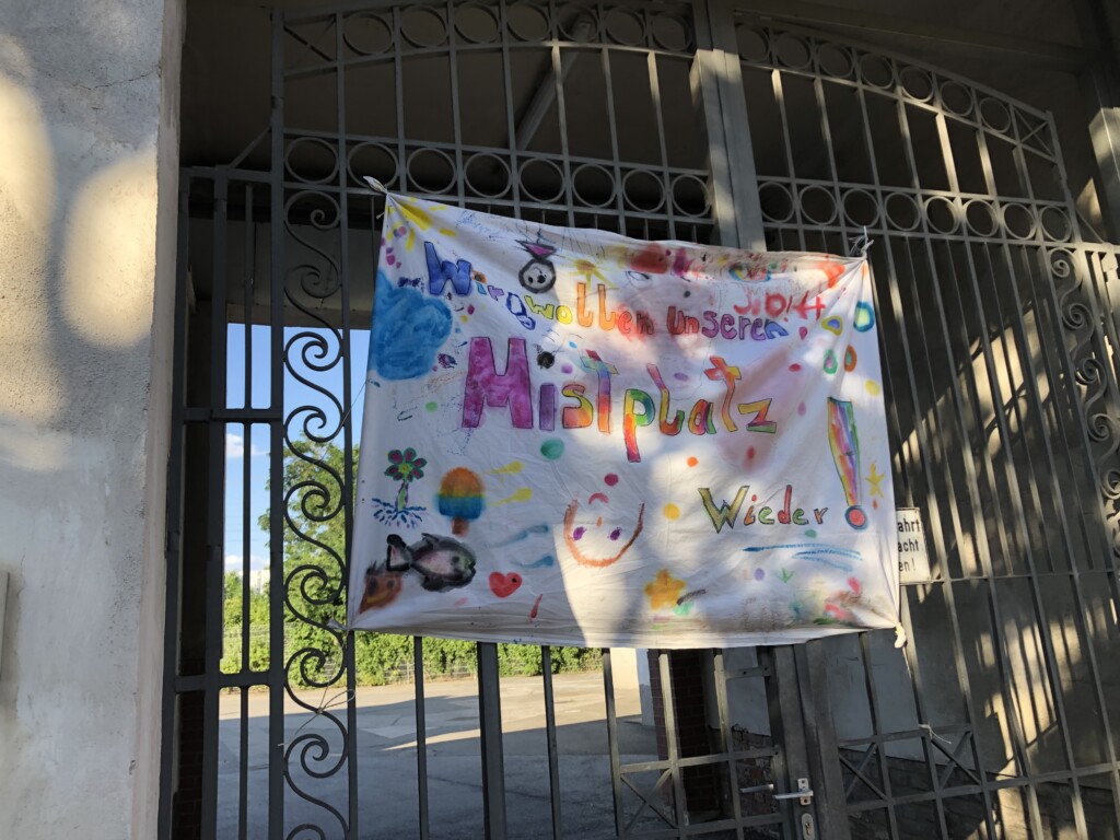 Transparent am Tor des alten Mistplatzes, auf dem in bunten Farben gemalt steht: "Wir wollen unseren Mistplatz wieder"