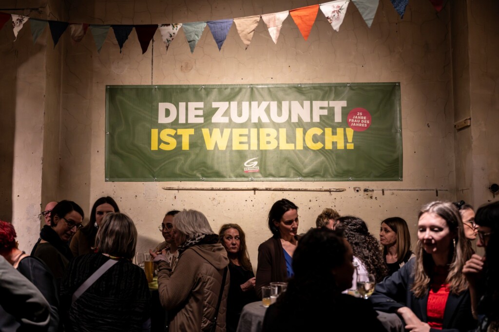 Gäste am Fest an Tischen plaudernd, dahinter ein Transparent: Die Zukunft ist weiblich.