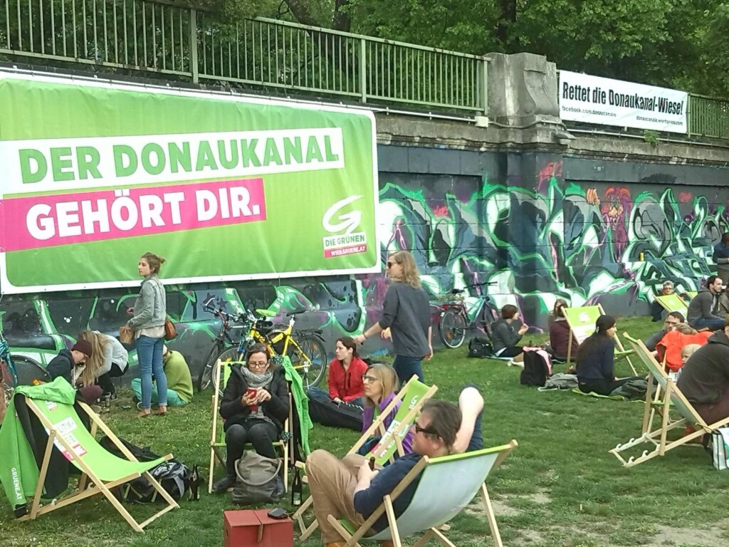 Menschen auf Liegestühlen, in einer Wiese sitzend, im Hintergrund ein Plakat: "Der Donaukanal gehört Dir."